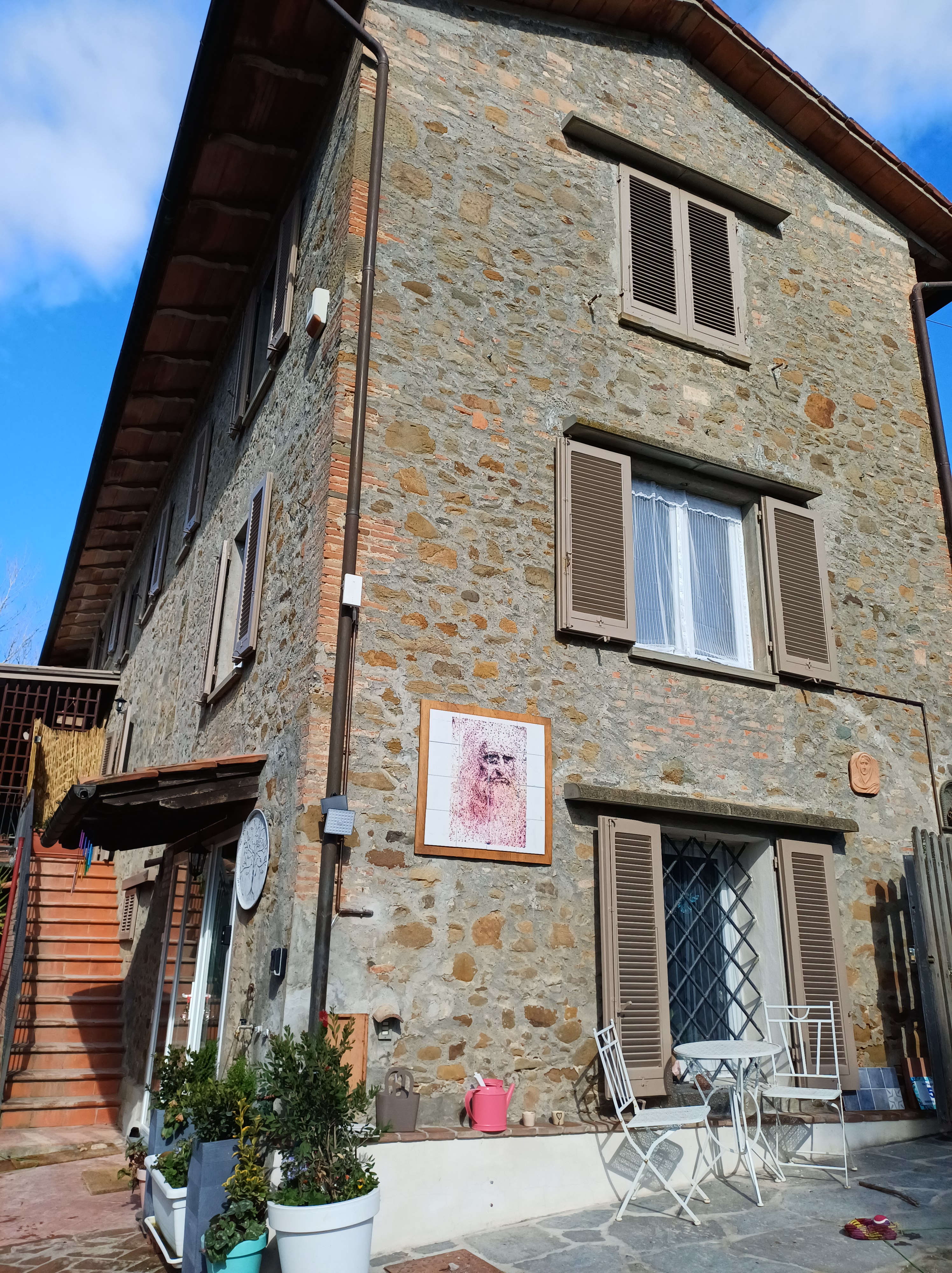 Rustic Italian stone house - Genevieve Grove, with wooden shutters, outdoor patio table and chairs, potted plants, and a framed artwork on the exterior wall.