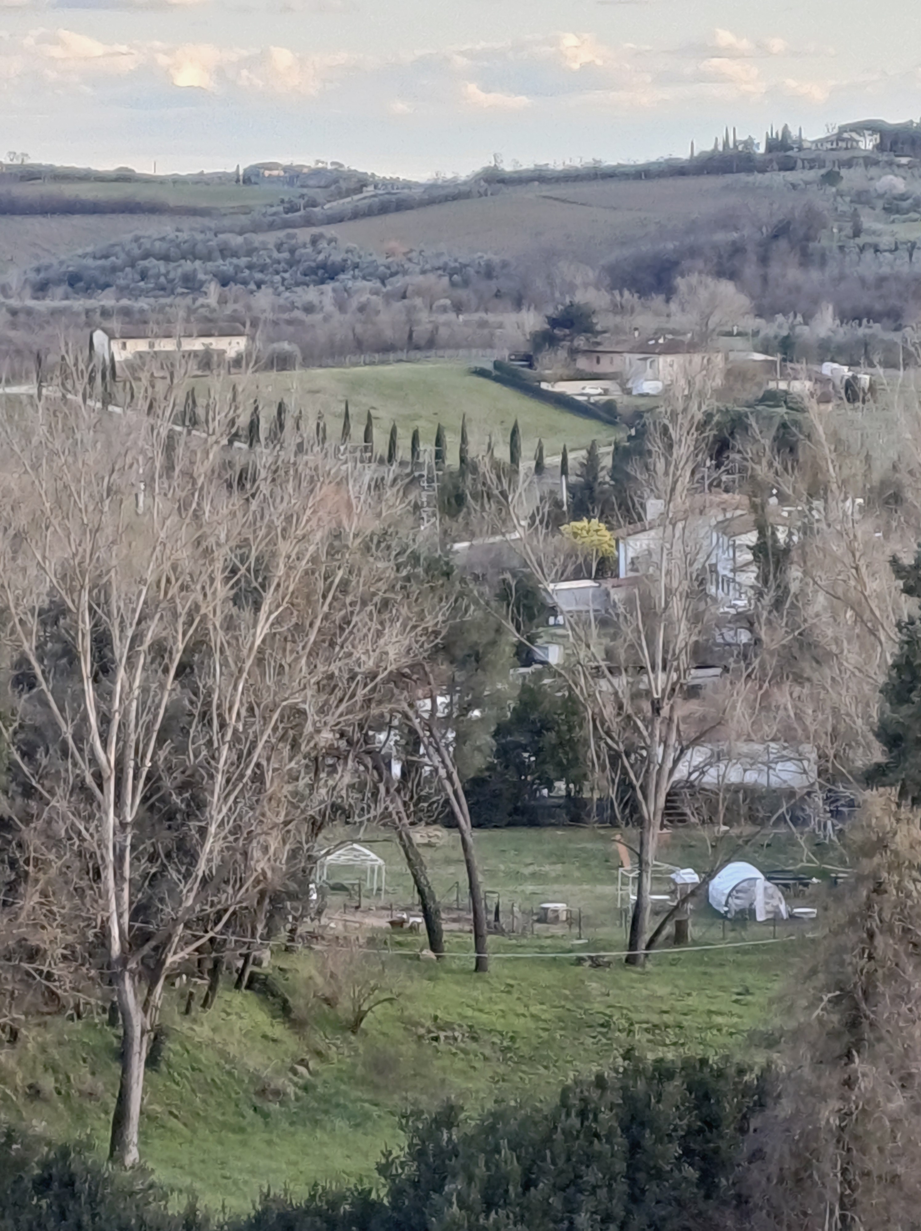 View of Genevieve Grove homestead from Vinci town overlooking Tuscan hills, farmland, cypress trees, and the rural garden below.